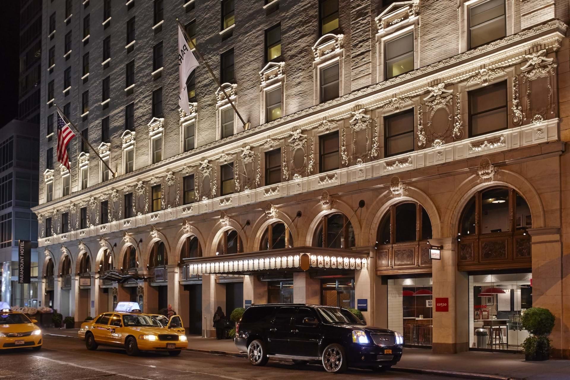 A nighttime view of a building's facade with arches, columns, and ornate details, with a street and cars in front.