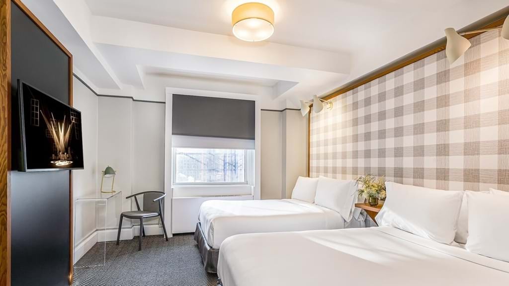 A hotel room with two beds, a TV, a window, a desk, and flowers.